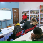 Aula della scuola di italiano di Cittadini del Mondo durante una lezione con LIM. L'aula è all'interno della sede dell'associazione, con librerie piene di libri sullo sfondo e un'atmosfera accogliente e inclusiva.